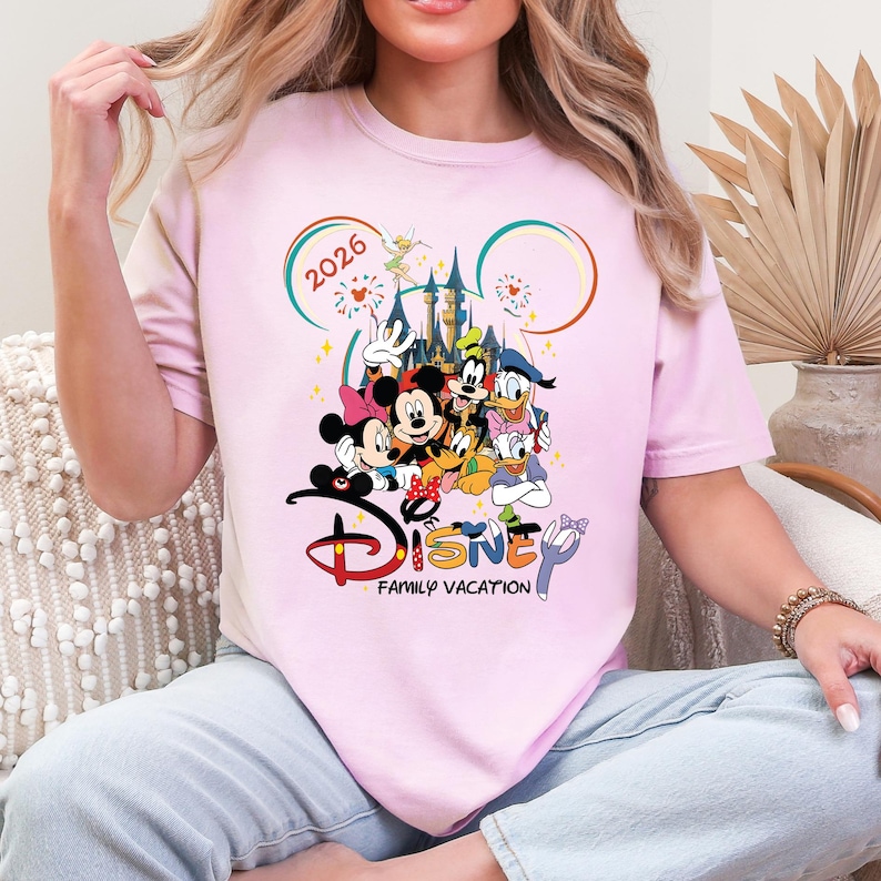 May include: A pale pink t-shirt with a Disney family vacation design. The graphic features Mickey and Minnie Mouse, Donald Duck, Goofy, and other characters in front of a castle. The text "2026" and "Disney Family Vacation" are also visible.