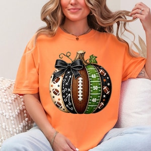 May include: An orange t-shirt featuring a graphic of a pumpkin designed with football elements. The pumpkin includes a black bow, a brown football texture with laces, a green field section with yard lines, and black panels with stars and plays.