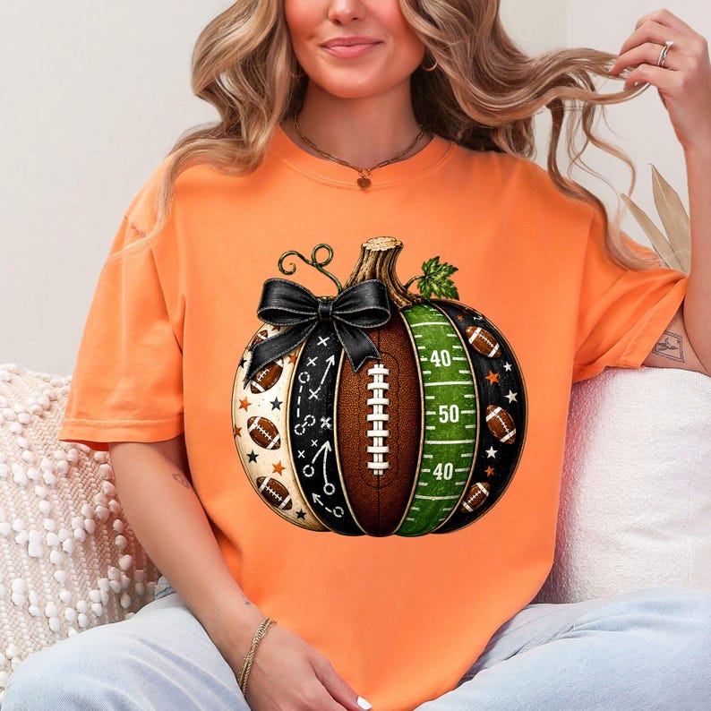 May include: An orange t-shirt featuring a graphic of a pumpkin designed with football elements. The pumpkin includes a black bow, a brown football texture with laces, a green field section with yard lines, and black panels with stars and plays.