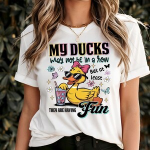 May include: White t-shirt with a graphic of a yellow duck wearing sunglasses and a pink bow, holding a drink. The text reads "MY DUCKS may not be in a row But at least THEY ARE HAVING Fun". The design includes butterflies and flowers.