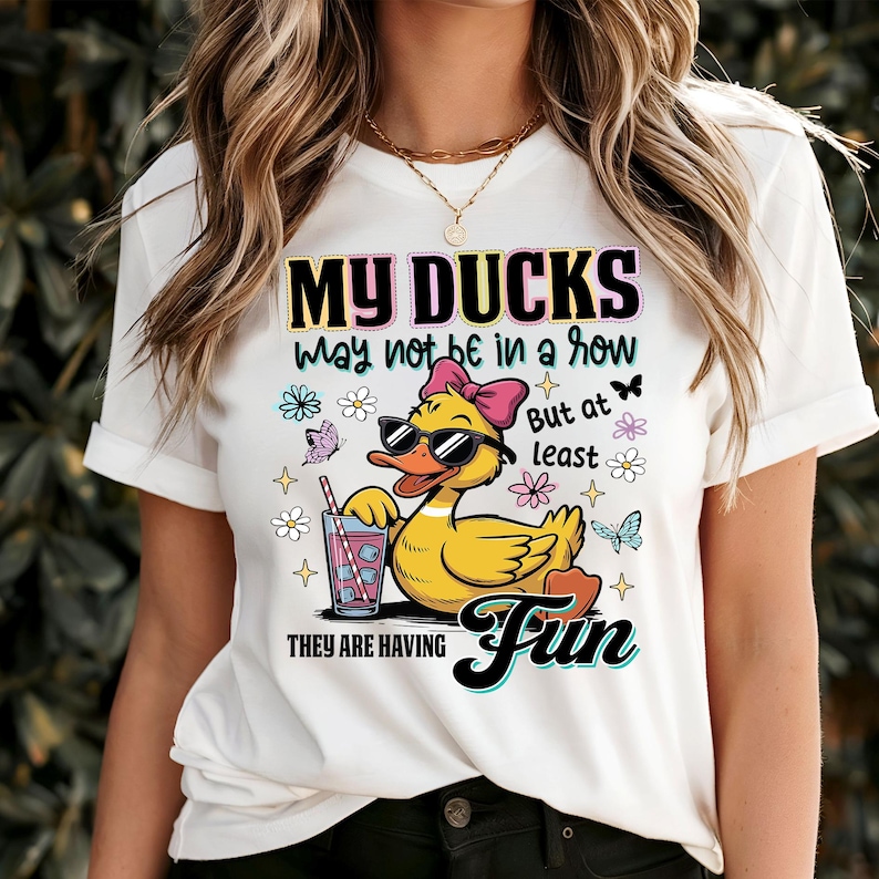 May include: White t-shirt with a graphic of a yellow duck wearing sunglasses and a pink bow, holding a drink. The text reads "MY DUCKS may not be in a row But at least THEY ARE HAVING Fun". The design includes butterflies and flowers.