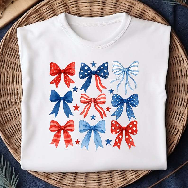 May include: White t-shirt featuring a watercolor design of nine bows in red, white, and blue. The bows are decorated with polka dots, stripes, and stars. The design is arranged in a grid pattern, with small stars scattered around the bows.