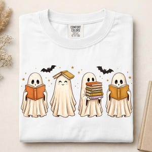 May include: A white t-shirt featuring a graphic of four cartoon ghosts reading books. The ghosts are off-white with black eyes and mouths. One ghost has a book on its head, another holds a stack of five books, and two hold open books. Small black bats and gold stars are scattered around the ghosts. The shirt has a Comfort Colors brand label.