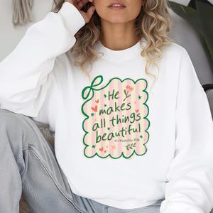May include: White sweatshirt with a colorful graphic that reads "He makes all things beautiful" in a whimsical font. The design includes pink stripes, hearts, flowers, and the Bible verse Ecclesiastes 3:11. A cozy and inspirational apparel item.