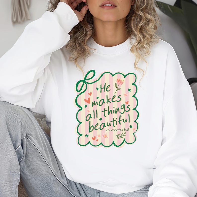 May include: White sweatshirt with a colorful graphic that reads "He makes all things beautiful" in a whimsical font. The design includes pink stripes, hearts, flowers, and the Bible verse Ecclesiastes 3:11. A cozy and inspirational apparel item.