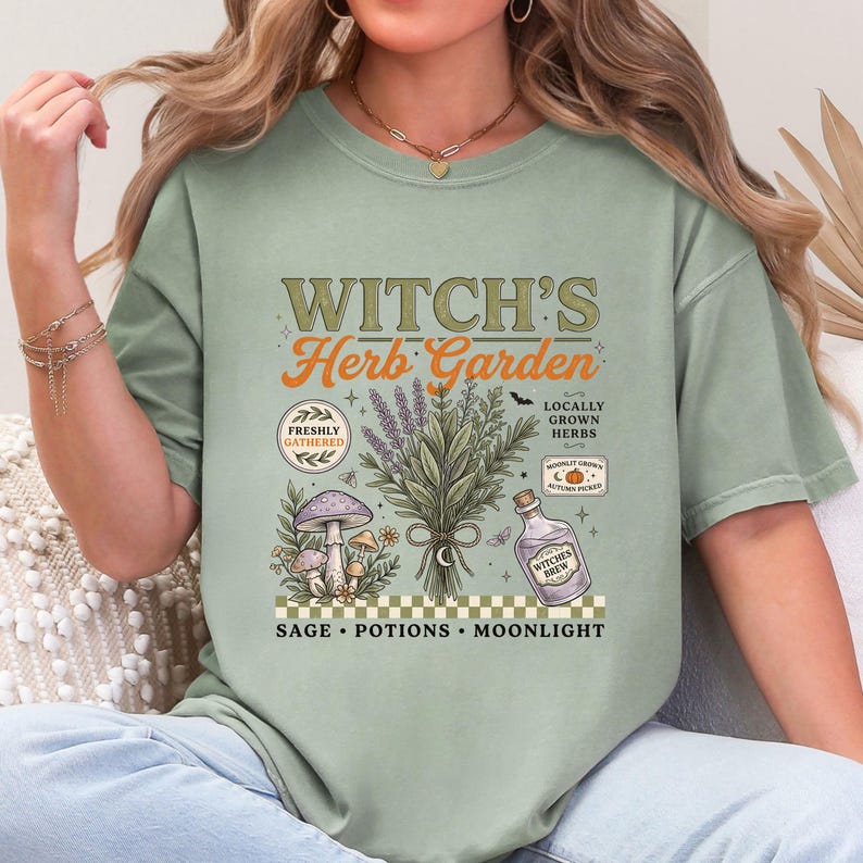May include: A sage green t-shirt featuring a graphic print with the text "WITCH'S Herb Garden" and "SAGE &bull; POTIONS &bull; MOONLIGHT". The design includes a central bundle of herbs, mushrooms, a potion bottle labeled "WITCHES BREW", and small icons.