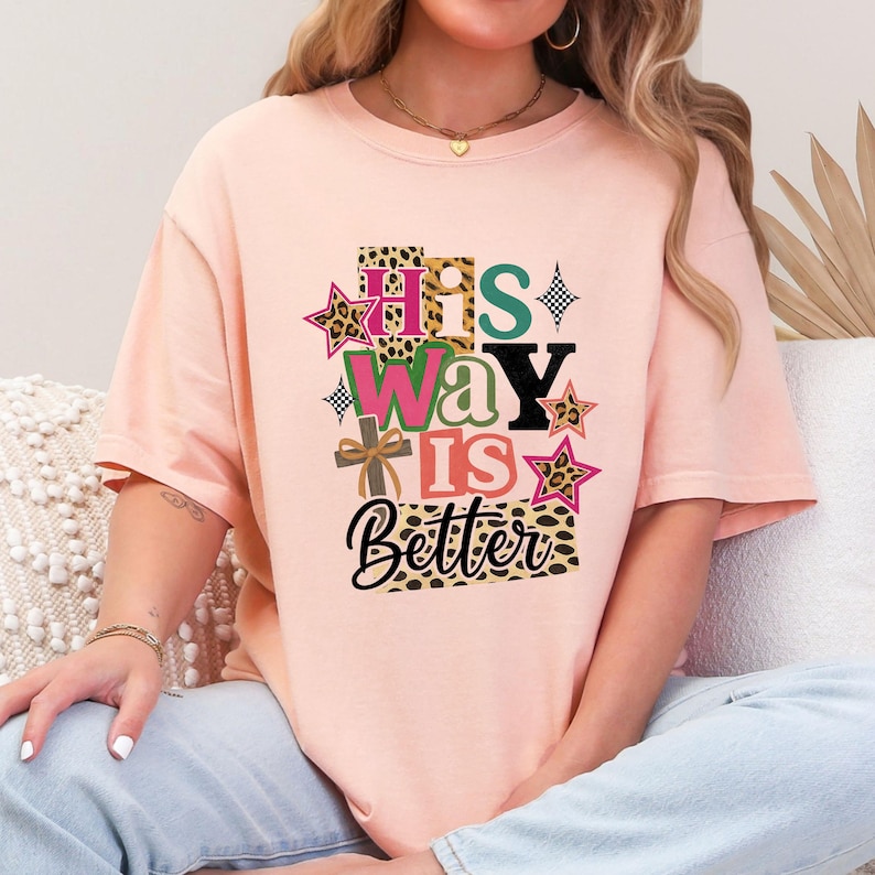 Op de afbeelding: Een licht perzikkleurig T-shirt met de tekst "HIS WAY IS Better" in een kleurrijk, gelaagd ontwerp. Het ontwerp bevat luipaardprint, sterren en een kruis. Het shirt is een casual, comfortabele stijl.