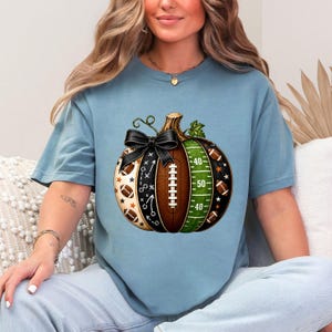 May include: A blue short-sleeve t-shirt featuring a graphic of a pumpkin divided into sections. The design includes football-themed patterns, a brown football texture, a green field with yard lines, and a black bow tied at the top stem.