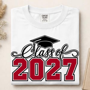 May include: A white short-sleeved t-shirt featuring a black graphic of a graduation cap above the black cursive text Class of and the large bold red numbers 2027. The shirt has a Comfort Colors brand label printed on the inside of the neck collar.