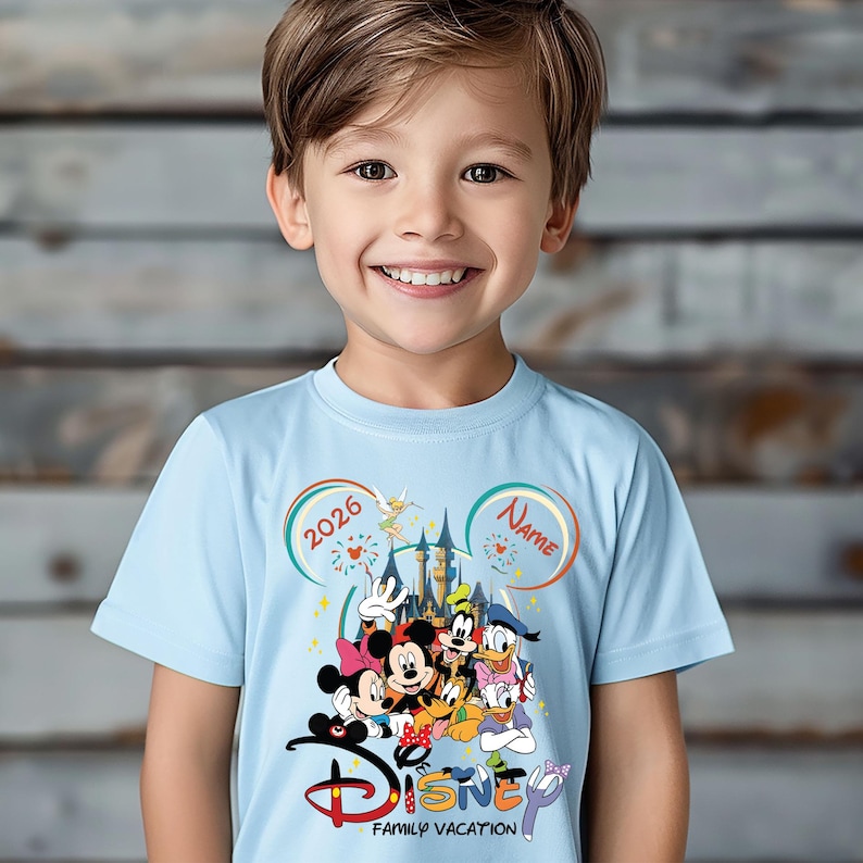 May include: A light blue t-shirt featuring a Disney Family Vacation design. The graphic includes Disney characters, a castle, fireworks, and the text "2026" and "Name" in colorful circles. The word "Disney" is in a stylized font.