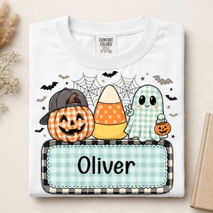 May include: A white t-shirt featuring a graphic of a pumpkin wearing a baseball cap, a piece of candy corn, and a ghost holding a small pumpkin bucket. Below the characters is a light blue gingham rectangle with the name Oliver printed in black.