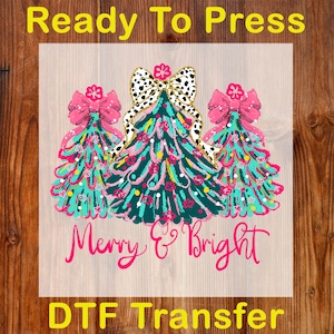 May include: A DTF transfer featuring three Christmas trees with pink bows and a black and white bow. The trees are teal and decorated with pink and yellow accents. The words "Merry & Bright" are written in pink script.
