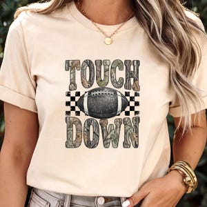 Pode incluir: Uma t-shirt de cor creme com uma impress&atilde;o gr&aacute;fica com o texto TOUCH DOWN num padr&atilde;o de camuflagem. Entre as palavras encontra-se uma ilustra&ccedil;&atilde;o de uma bola de futebol americano a preto e branco sobre um fundo axadrezado.