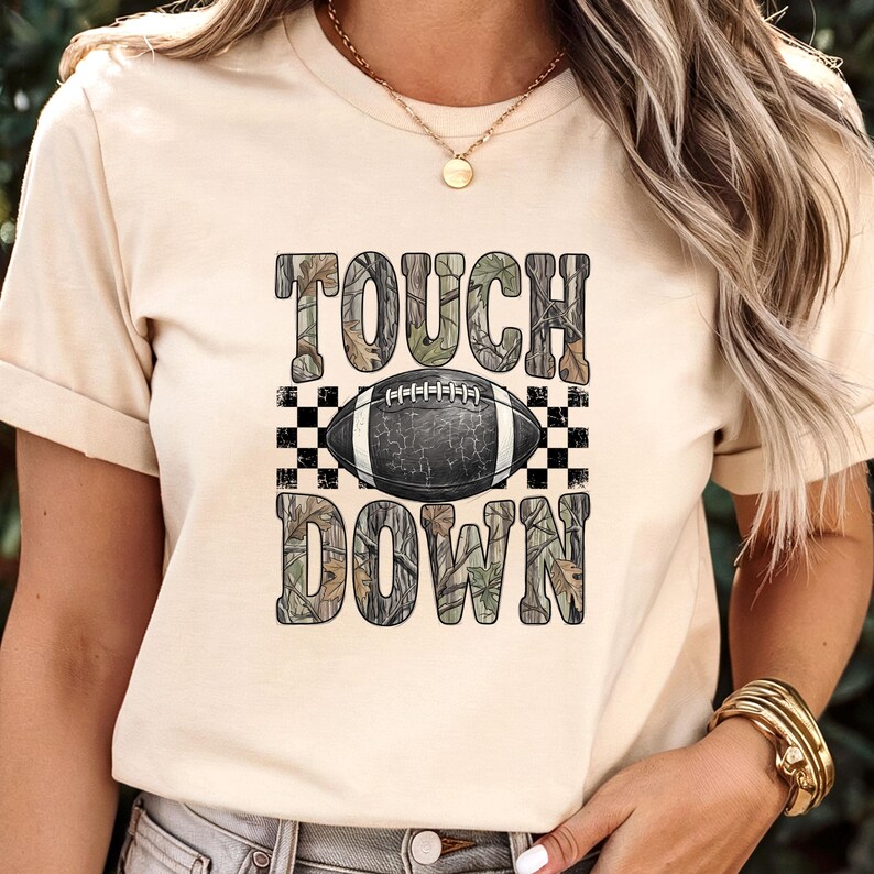 Pode incluir: Uma t-shirt de cor creme com uma impress&atilde;o gr&aacute;fica com o texto TOUCH DOWN num padr&atilde;o de camuflagem. Entre as palavras encontra-se uma ilustra&ccedil;&atilde;o de uma bola de futebol americano a preto e branco sobre um fundo axadrezado.