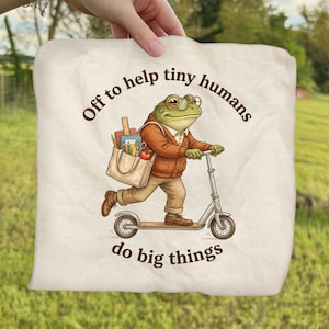 May include: Off-white tote bag featuring a cartoon frog riding a scooter. The frog wears glasses, a sweater, and carries a tote bag filled with school supplies. Text on the bag reads "Off to help tiny humans do big things."
