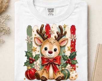 Könnte beinhalten: Ein weißes T-Shirt mit einer Grafik eines braunen Rentiers mit Geweih, das eine große rote Schleife trägt. Das Design zeigt grüne Stechpalmenblätter mit roten Beeren, goldene Sterne, weiße Schneeflocken sowie rote und grüne Kugeln vor einem Streifenhintergrund.