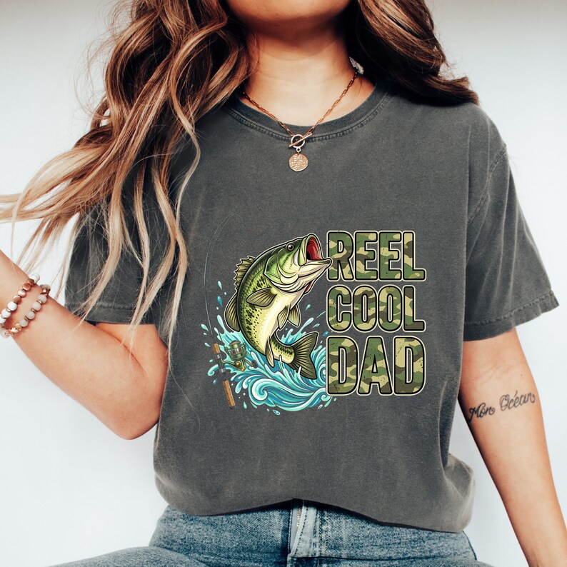 Pu&ograve; includere: T-shirt grigio scuro con una grafica di un pesce persico che salta e la scritta "REEL COOL DAD" in mimetica. Il design include schizzi d'acqua e attrezzatura da pesca, creando un design a tema pesca.