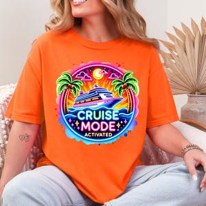May include: Orange t-shirt featuring a vibrant graphic with a cruise ship, palm trees, and the text "CRUISE MODE ACTIVATED" in a circular design. The design includes blue, pink, and yellow accents, creating a tropical vacation theme.