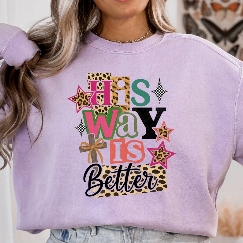 Op de afbeelding: Een lichtpaarse sweatshirt met de tekst "His Way Is Better" in een mix van lettertypen en kleuren. Het ontwerp bevat luipaardprint, sterren en een kruis. De sweatshirt is een casual, comfortabele stijl.