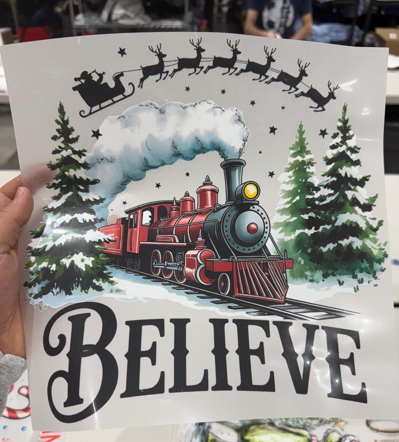 Może przedstawiać: Świąteczna grafika przedstawiająca czerwony pociąg parowy, dym i sanie ciągnięte przez renifery. Słowo &bdquo;BELIEVE&rdquo; jest napisane dużymi czarnymi literami poniżej. Obraz zawiera choinki i zaśnieżony krajobraz.