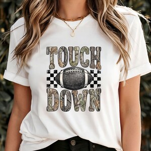 Pode incluir: Uma t-shirt branca de manga curta com uma impress&atilde;o gr&aacute;fica com o texto TOUCH DOWN num padr&atilde;o de camuflagem. Uma bola de futebol americano preta e branca est&aacute; centrada entre as palavras, ladeada por quadrados axadrezados pretos e brancos.