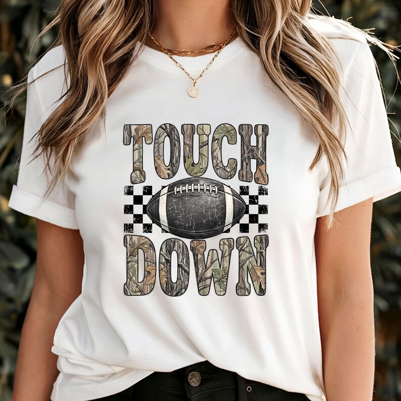 Pode incluir: Uma t-shirt branca de manga curta com uma impress&atilde;o gr&aacute;fica com o texto TOUCH DOWN num padr&atilde;o de camuflagem. Uma bola de futebol americano preta e branca est&aacute; centrada entre as palavras, ladeada por quadrados axadrezados pretos e brancos.
