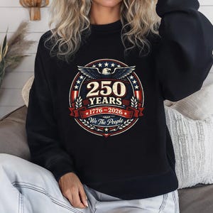 Peut inclure: Sweat-shirt noir avec un motif circulaire comm&eacute;morant 250 ans. Le design comprend un aigle, des &eacute;toiles et le texte "250 YEARS 1776-2026 We The People" en rouge, blanc et bleu.