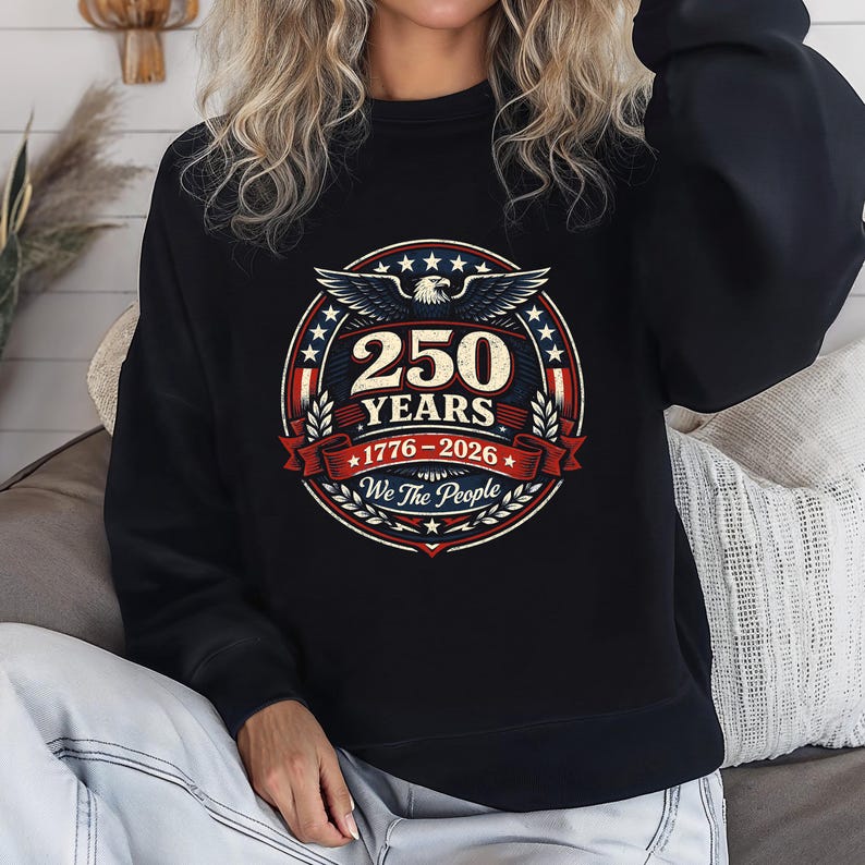 Peut inclure: Sweat-shirt noir avec un motif circulaire comm&eacute;morant 250 ans. Le design comprend un aigle, des &eacute;toiles et le texte "250 YEARS 1776-2026 We The People" en rouge, blanc et bleu.