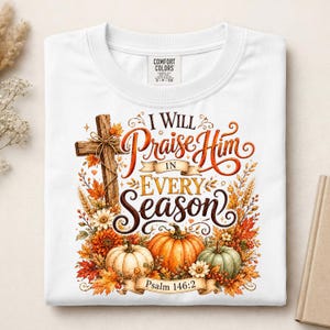 May include: A white t-shirt featuring a graphic print with a wooden cross, orange and green pumpkins, autumn leaves, and flowers. The text reads I WILL Praise Him IN EVERY Season and Psalm 146:2. The shirt has a Comfort Colors brand neck label.
