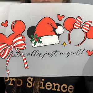 Peut inclure: Une feuille transparente avec des motifs rouges et blancs sur le th&egrave;me de Mickey Mouse. Comprend un n&oelig;ud, un bonnet de P&egrave;re No&euml;l et des oreilles de Minnie Mouse. Le texte "Literally just a girl!" est &eacute;crit en cursive. Une &eacute;toile jaune est &eacute;galement pr&eacute;sente.