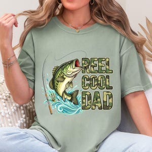 Pu&ograve; includere: T-shirt verde salvia con grafica di un persico trota che salta fuori dall'acqua, una canna da pesca e la scritta "REEL COOL DAD" in camouflage. La maglietta ha una vestibilit&agrave; ampia.