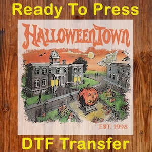 May include: A DTF transfer featuring a Halloween Town design. The design includes a spooky town scene with a jack-o'-lantern, buildings, and the text "HALLOWEENTOWN" and "EST. 1998". The words "Ready To Press" and "DTF Transfer" are also present.