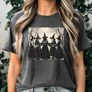 Puede incluir: Camiseta gris oscuro con un gr&aacute;fico en blanco y negro de seis brujas con vestidos largos y sombreros puntiagudos. El texto "Girls Will Be Girls" est&aacute; impreso encima de las brujas. La camiseta tiene mangas cortas y cuello redondo.