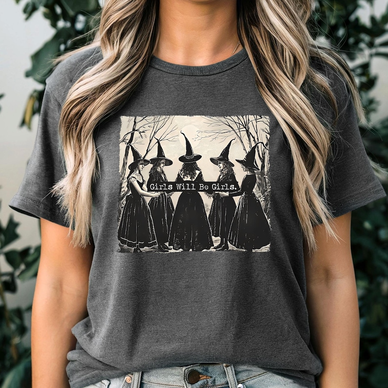Puede incluir: Camiseta gris oscuro con un gr&aacute;fico en blanco y negro de seis brujas con vestidos largos y sombreros puntiagudos. El texto "Girls Will Be Girls" est&aacute; impreso encima de las brujas. La camiseta tiene mangas cortas y cuello redondo.