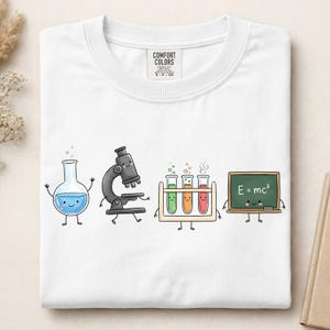 May include: A white t-shirt featuring a graphic print of four anthropomorphic science items: a blue liquid-filled flask, a black microscope, a wooden rack with three colorful test tubes, and a green chalkboard with the text E=mc2 written on it.