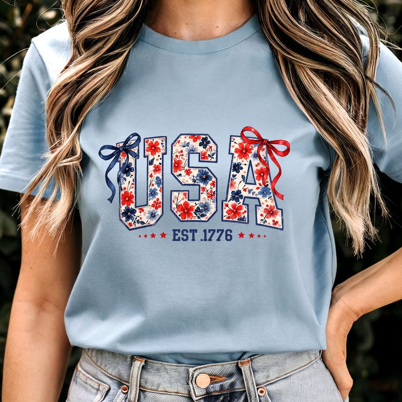 Może przedstawiać: Jasnoniebieska koszulka z napisem "USA" w kwiatowy wz&oacute;r z czerwonych i niebieskich kwiat&oacute;w. Tekst "EST. 1776" znajduje się pod słowem "USA" z gwiazdkami po każdej stronie. Koszulka ma kr&oacute;tkie rękawy.