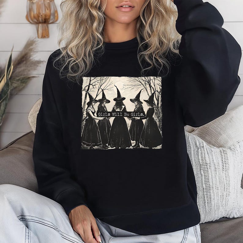 Puede incluir: Sudadera negra con un gr&aacute;fico de cinco brujas con vestidos y sombreros negros, sobre un fondo de bosque. El texto "Girls Will Be Girls" est&aacute; impreso en el gr&aacute;fico. Cuello redondo.