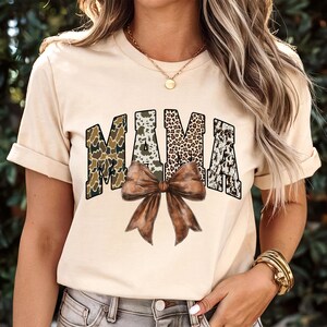 May include: A beige t-shirt with the word "MAMA" in a bold, block letter design. The letters feature camouflage, leopard print, and cowhide patterns. A large brown bow is centered below the word.