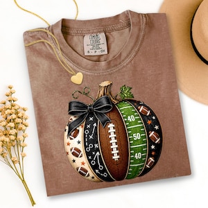 May include: A brown short-sleeve t-shirt featuring a graphic of a pumpkin designed with football elements, including a green field section with yard lines, a brown football texture, and a black bow. The shirt has a Comfort Colors brand label.
