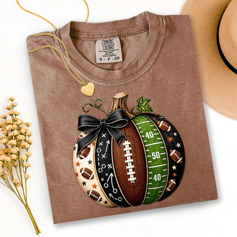 May include: A brown short-sleeve t-shirt featuring a graphic of a pumpkin designed with football elements, including a green field section with yard lines, a brown football texture, and a black bow. The shirt has a Comfort Colors brand label.