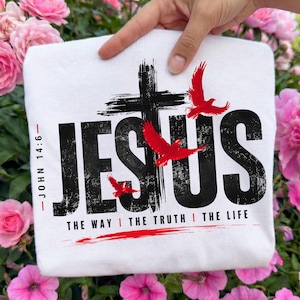 May include: A white t-shirt featuring a black distressed graphic of the word JESUS with a cross replacing the letter T. Three red birds are flying around the text. The text JOHN 14:6 and THE WAY THE TRUTH THE LIFE are printed in black.