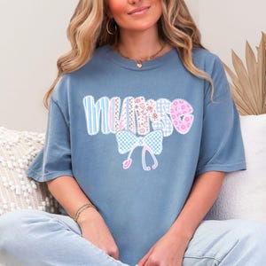 May include: A blue t-shirt with the word "NURSE" in a patchwork design. The letters are in various patterns, including stripes, gingham, and floral prints. A bow and stethoscope graphic is below the word.