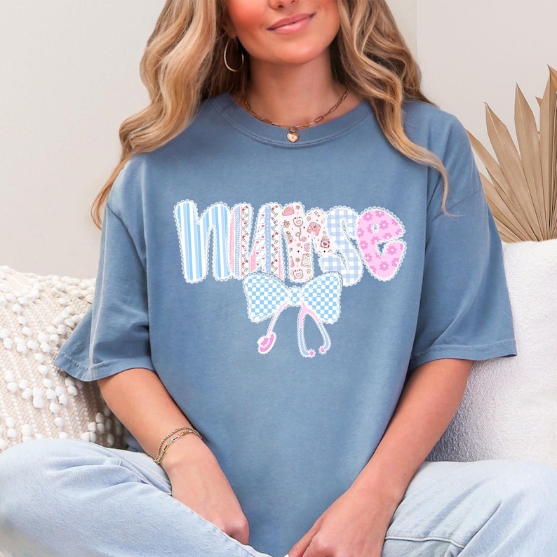 May include: A blue t-shirt with the word "NURSE" in a patchwork design. The letters are in various patterns, including stripes, gingham, and floral prints. A bow and stethoscope graphic is below the word.