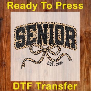 May include: A DTF transfer design with the word "SENIOR" in a black glitter font, outlined with a leopard print pattern. A leopard print bow is below the word, with "EST. 2026" in a smaller font. The words "Ready To Press" and "DTF Transfer" are also included.