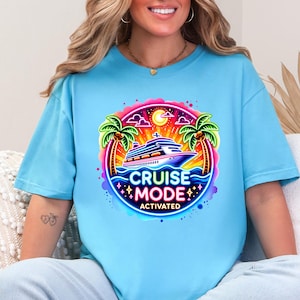 May include: Light blue t-shirt featuring a vibrant graphic with a cruise ship, palm trees, and the text "CRUISE MODE ACTIVATED." The design is framed by a colorful border with stars and a sun. A casual, comfortable tee.