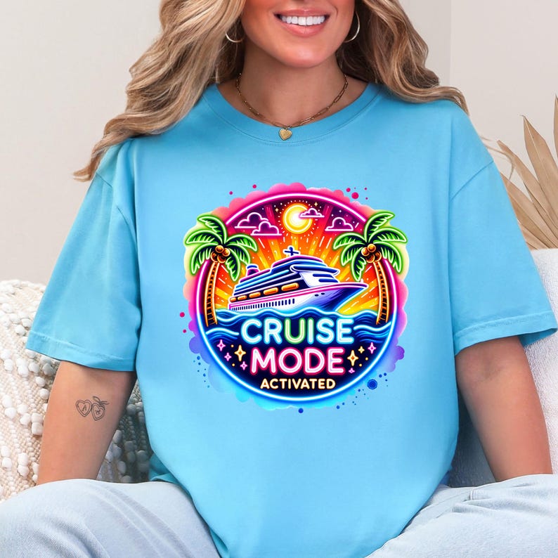 May include: Light blue t-shirt featuring a vibrant graphic with a cruise ship, palm trees, and the text "CRUISE MODE ACTIVATED." The design is framed by a colorful border with stars and a sun. A casual, comfortable tee.