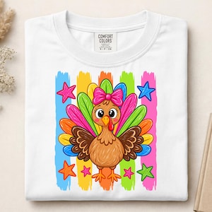 Puede incluir: Una camiseta blanca con un gráfico de un pavo marrón con un lazo rosa en la cabeza. El pavo está centrado sobre rayas verticales azules, amarillas, verdes y rosas, rodeado de estrellas coloridas. La etiqueta interior del cuello dice COMFORT COLORS.