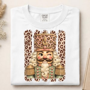 May include: A white t-shirt featuring a graphic of a winking nutcracker with a brown mustache and white beard. The nutcracker wears a leopard print hat and uniform accents, set against a vertical leopard print background. Comfort Colors label.