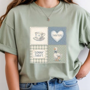 May include: Sage green t-shirt featuring a patchwork design with a teacup, heart, goose, and text that reads "Thrifters" and "Gonna Thrift". The design has a vintage, quilted aesthetic.