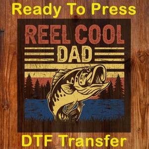 May include: A DTF transfer with a fishing theme. The design features the text "REEL COOL DAD" in red and yellow, with a fish illustration. The background includes a sunset and water scene. The words "Ready To Press" and "DTF Transfer" are also present.